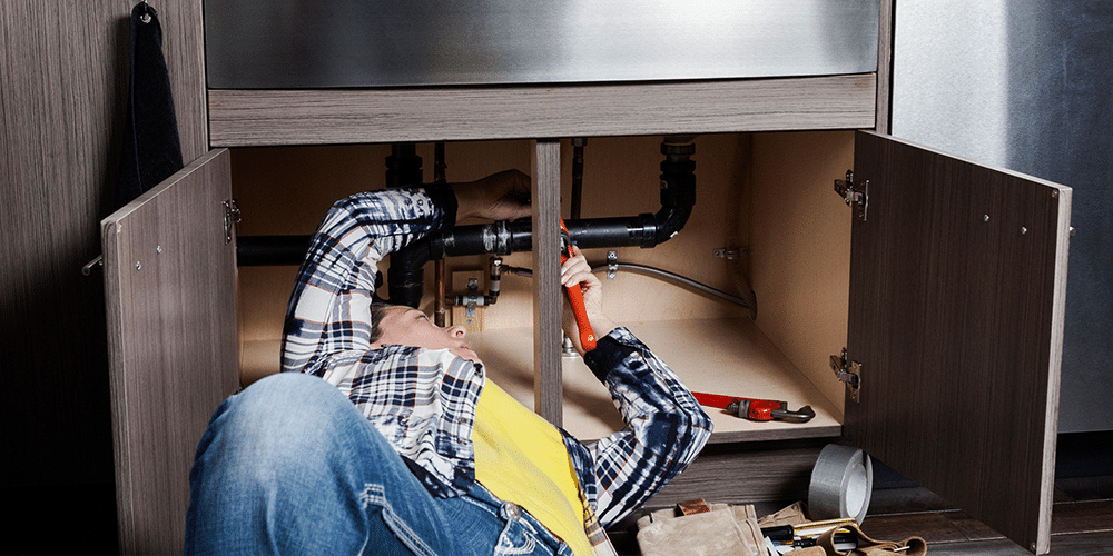 Woman in yellow shirt checks pipes underneath sink.
