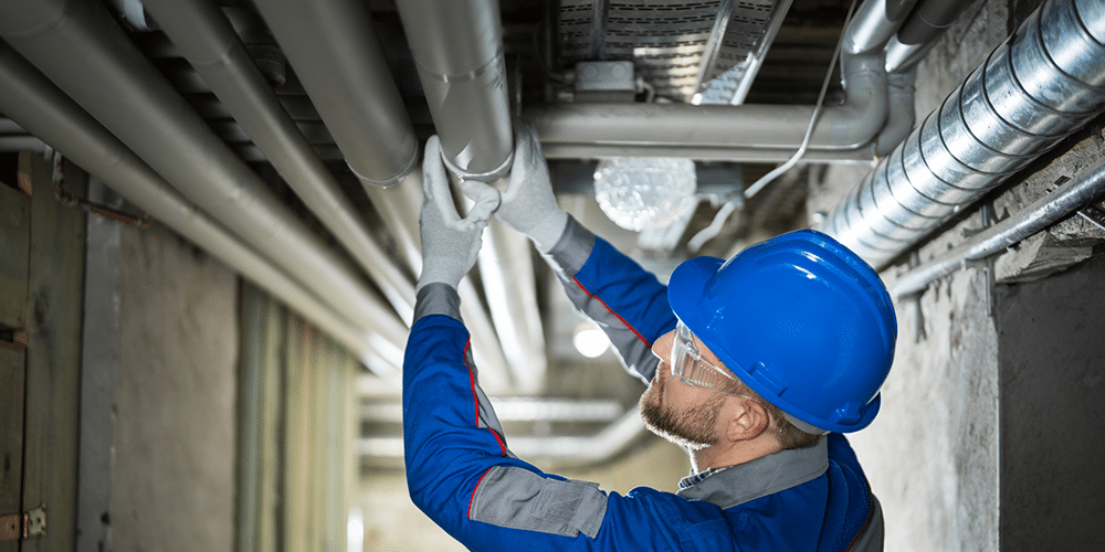 Technician in blue uniform and hardhat inspects pipes in basement.