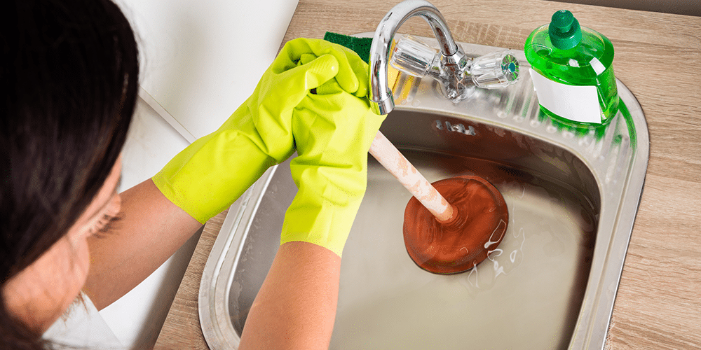 Woman wearing yellow kitchen gloves using a plunger to unclog a bathroom sink.
