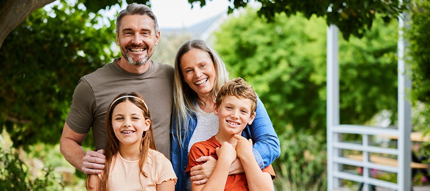 Mother, father, and two kids embrace on the back porch of their home with woods in the background.