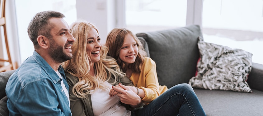 Mother, father, and daughter snuggle on couch and laugh at something on TV.