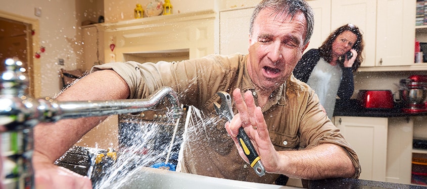 Male homeowner faces water from broken faucet while wife calls for help.