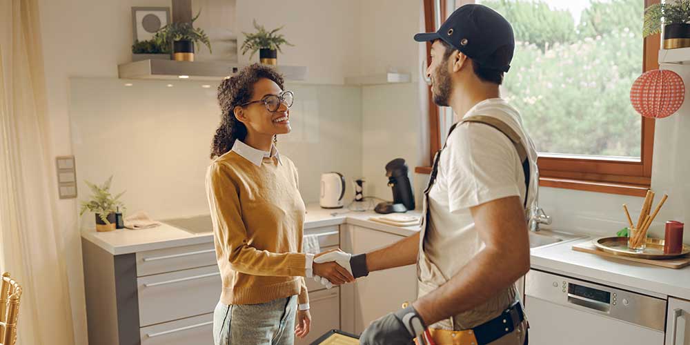 Young female homeowner smiles and shakes hands with a male local plumber in her kitchen.